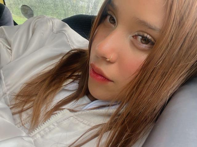 HeraJuno has light brown skin, long brown hair, and a nose ring. She's wearing a white jacket, looking at the camera with slightly parted pink lips.