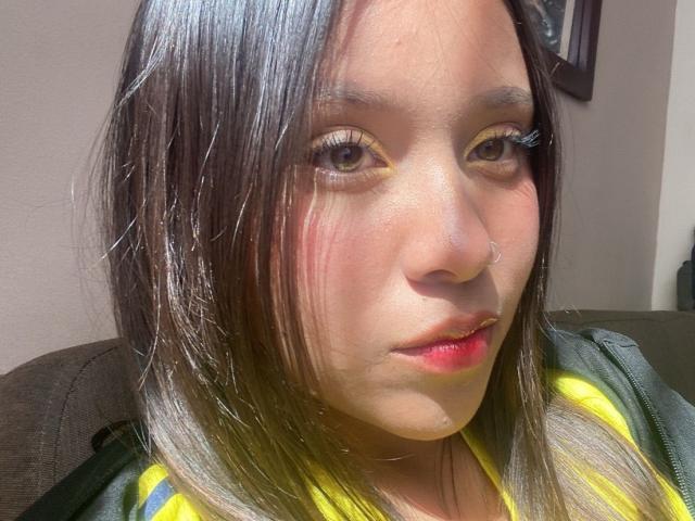 HeraJuno's face, with shiny brown hair, yellow eyeliner, and red lipstick, showing a slight smile, wearing a green and yellow striped shirt, sunlit.