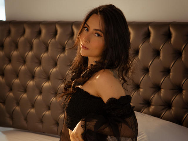 A photograph of HotFemme with long, wavy dark brown hair and light olive skin. She's wearing an off-shoulder black lace top, revealing a hint of her upper chest and shoulders. She gazes directly at the camera with a subtle, confident smile. The tufted, dark brown headboard in the background contrasts with her light skin and dark hair. Soft, natural light enhances her facial features and the texture of her lace top. She's positioned slightly to the right, with her left arm resting near her chest. The composition emphasizes her relaxed yet poised demeanor.