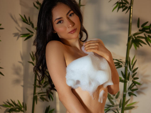 A photograph of a HotFemme with long, dark brown hair and light olive skin. She's standing with green bamboo plants in the background, holding a foamy white substance that covers her small to medium-sized breasts. Her brown eyes are slightly downcast, and she has a subtle, confident smile. The soft lighting highlights her smooth skin and natural beauty. The foamy substance contrasts with her skin tone and adds a sense of playful innocence to the image. The bamboo plants add a natural, serene element to the composition.