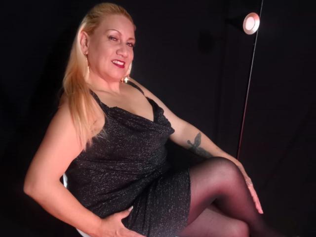 MatureYayita with blonde hair, red lipstick, and sparkly black dress, sitting, showing her ass, tattoo on right arm, smiling, legs crossed, under soft light.
