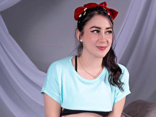 LeahMuniz with dark hair in a red bow, wearing a light blue crop top, black bra, and earbuds, smiling slightly, showing a cute face and small tits.