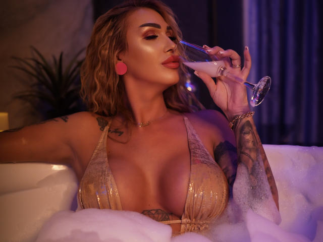 NicolSNaya, con cabello rubio ondulado, audífonos rosas y una blusa beige brillante de escote bajo, bebiendo un martini, mostrando sus grandes senos y tatuajes, en una bañera espumosa..