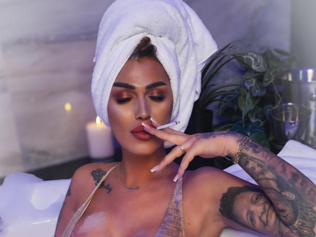 NicolSNaya met een witte handdoek op haar hoofd, tatoeages op haar arm en haar hand in de buurt van haar lippen, draagt een doorschijnende top en zit in een bad met een kaars..