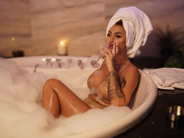 NicolSNaya with a towel on her head, naked, in a bubbly white tub, holding a glass of wine, tattoo on her arm, sucking on a red lollipop, tan skin.