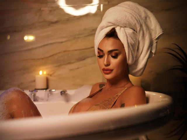 NicolSNaya in a bathtub, her tan skin glowing, white towel wrapped around her head, small breasts partially visible, candlelit, water bubbles on her skin, looking down with a calm expression.