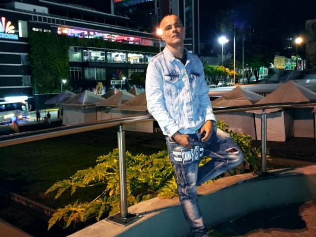 StrongAlan leans against a metal railing, wearing a light blue denim jacket with patches and ripped blue jeans. He has short, dark hair and a serious expression. The background is a brightly lit urban area with buildings and neon signs. StrongAlan's right leg is bent at the knee, resting on the railing. The image uses natural and artificial lighting, highlighting StrongAlan's casual, relaxed stance. The texture of the denim and metal railing is clearly detailed.
