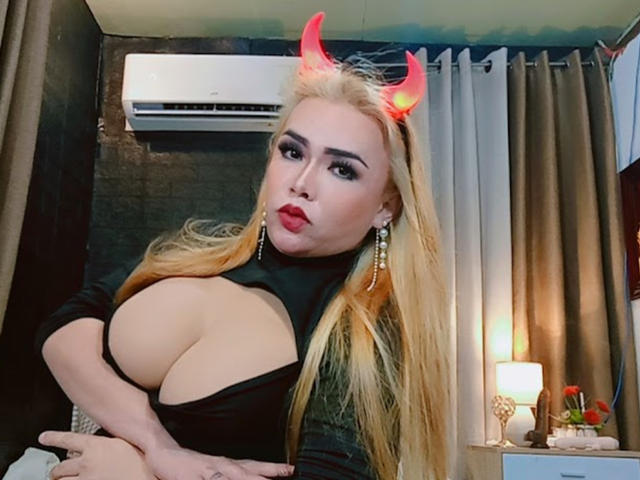 NathalieHotCocx with blonde hair, red devil horns, and a black cut-out top showing her big tits. She's looking at the camera with red lipstick and earrings.