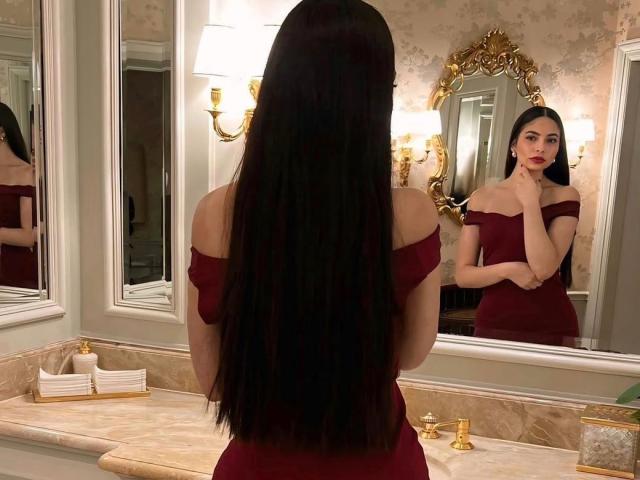 LolaHeaven with long black hair, off-shoulder red dress, stands in front of mirror, touching her red lips, reflection shows her serious expression, elegant bathroom setting.