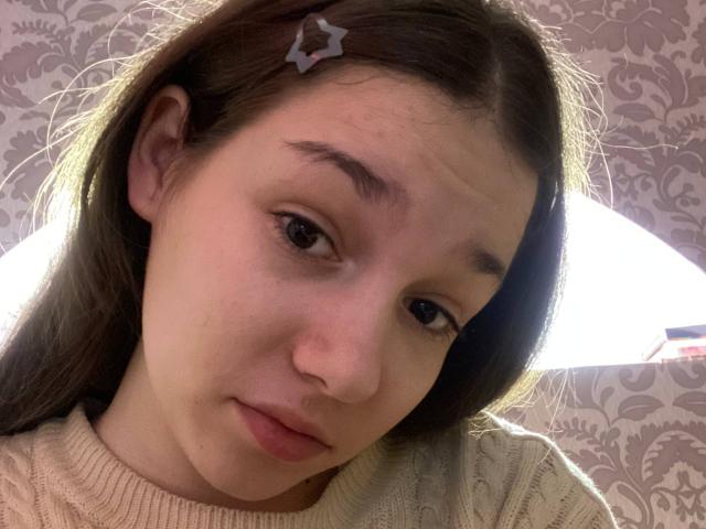 KaiaLein a les cheveux bruns et porte une pince à cheveux en forme d'étoile. Elle est vêtue d'un pull blanc en tricot, regarde légèrement sur le côté avec une expression neutre, et a une petite imperfection sur sa joue..