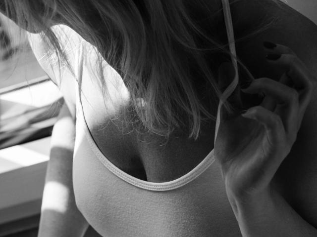 Gerdaonly-hot's tits are visible in a black-and-white photo, wearing a white, thin-strapped, low-cut top, with sunlight casting shadows on her chest and hand near her neck.