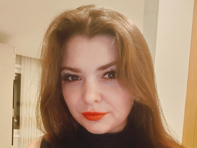 CherryMagdalena a du rouge à lèvres, de longs cheveux bruns, et porte du maquillage noir avec de longs cils. Son nez est légèrement retroussé. Elle regarde directement l'appareil photo..