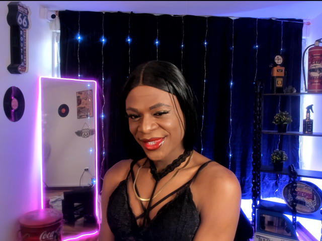 VenusMiller with black hair, red lipstick, and black lace top, smiling at camera, showing her tits in a room with blue curtains and string lights.