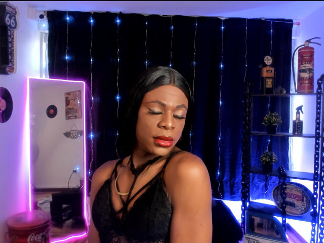 VenusMiller stands in a room with black curtains and blue string lights. She has dark skin, straight black hair, and wears a black lace strappy top. Her eyes are closed, and she has red lipstick. Neon pink light on the left and shelves with figurines on the right. Her makeup is bold, with defined eyebrows and eyeliner. She has a confident, relaxed expression. The lighting highlights her facial features and hair. The image has a modern, slightly edgy vibe.