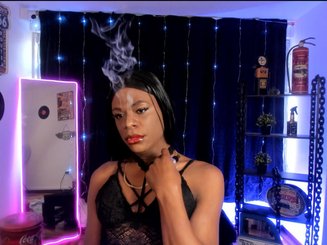 Venus Miller, con un taglio di capelli corto neri, che fuma, indossa una camicetta di pizzo nero, rossetto rosso, tiene il colletto, montatura rosa neon, tenda nera, luci blu, mensola con statuine e piante. Il seno è visibile..
