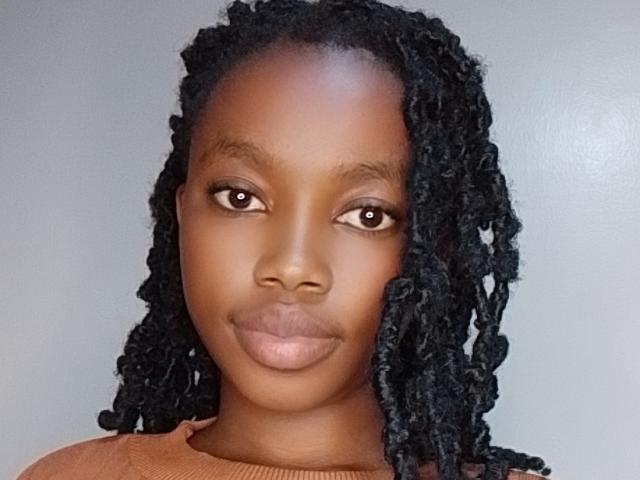 CutyLipsy has dark brown skin, straight black dreadlocks, brown eyes, and full pink lips, wearing a brown sweater, looking straight at the camera with a neutral expression.