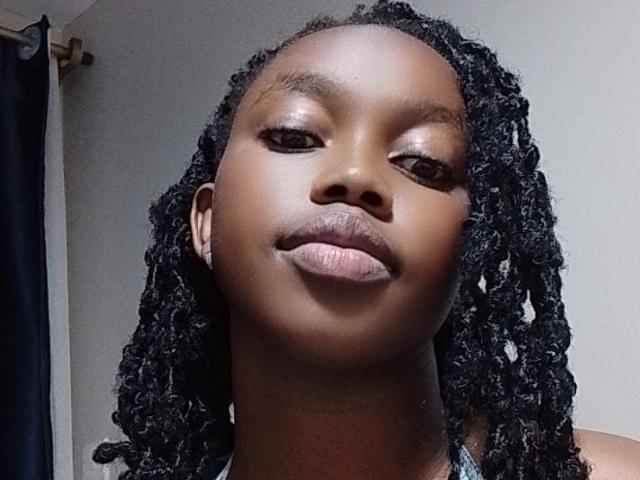 CutyLipsy, con piel marrón oscuro, rastas rizadas negras y labios rosados y gruesos, mirando ligeramente hacia abajo con una sonrisa sutil. Su rostro está centrado, con un fondo neutro..