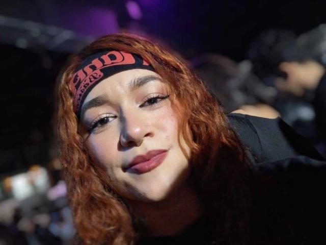 Valery Hilton, con cabello rojo ondulado, una diadema negra, un piercing en la nariz y lápiz labial oscuro, sonriendo ligeramente, vestida de negro, con un fondo borroso y luz púrpura. No se ven los senos..