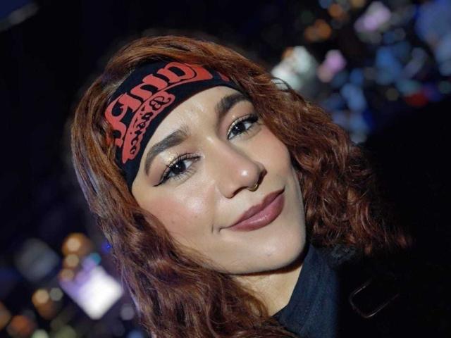 Valery Hilton, con cabello castaño ondulado, lleva una diadema negra con letras rojas, sonríe con los labios rosados ligeramente entreabiertos, delineador de ojos oscuro y una chaqueta oscura en una noche de ciudad..