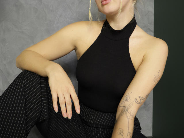 SummerBirch met bleke huid, blond haar, een zwarte haltertop, een zwart rokje met streepjes, een getatoeëerde arm, zittend, met blote schouders, armtattoo, ontspannen, lichte glimlach, hand op de knie..