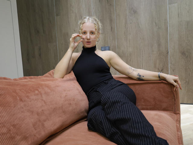 SummerBirch, con i capelli biondi ricci, indossa un top nero a halter e pantaloni neri a righe e siede su un divano rosso in velluto, mostrando dei tatuaggi sul braccio destro..