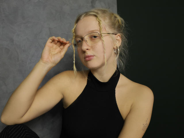 SummerBirch, avec ses tresses blondes, ses lunettes et son haut noir à encolure dégagée, ajustant ses lunettes, montrant un tatouage sur son bras droit, et une expression confiante, debout devant un fond sombre..