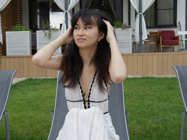 SheilaPass sits on a gray lounge chair, hands in her long, wavy black hair. She wears a white, deep V-neck dress with black lace trim. Her light skin contrasts with the green grass. The background includes a wooden deck, white curtains, and red chairs. The image has a natural, candid feel. Her expression is calm, with slight curiosity. She has small earrings. The texture of the dress is knit.
