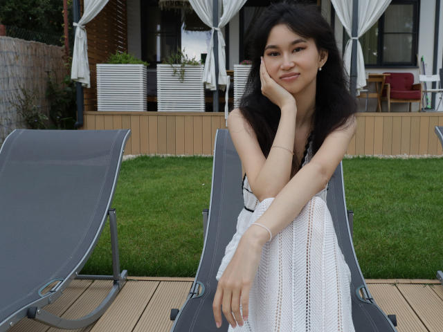 SheilaPass with long black hair, sitting on a gray lounge chair, wearing a white dress, hand on cheek, smiling, grassy backyard, wooden deck, white curtains in background.