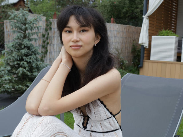 SheilaPass with dark hair, cute face, small tits, sitting on gray chair, wearing white lace dress, hands on neck, outdoors, wooden fence, green tree, white umbrella in background.