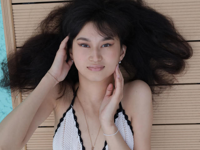 SheilaPass has medium brown, slightly wavy hair that's spread out around her head. She's looking at the camera with a subtle, confident smile. Her hands are gently touching her face, with her right hand on her cheek and her left hand on her temple. She's wearing a white crochet halter top with black trim and small black beads. She has light skin, brown eyes, and is wearing small stud earrings. A thin silver bracelet is on her right wrist. The light in the photo is even, highlighting her facial features and hair texture. The image is sharp and detailed.
