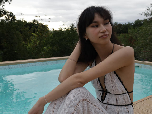 SheilaPass a de petites seins, assise au bord d'une piscine, vêtue d'une robe blanche en tricot avec des détails noirs, regardant sur le côté, avec des cheveux foncés et un léger sourire..
