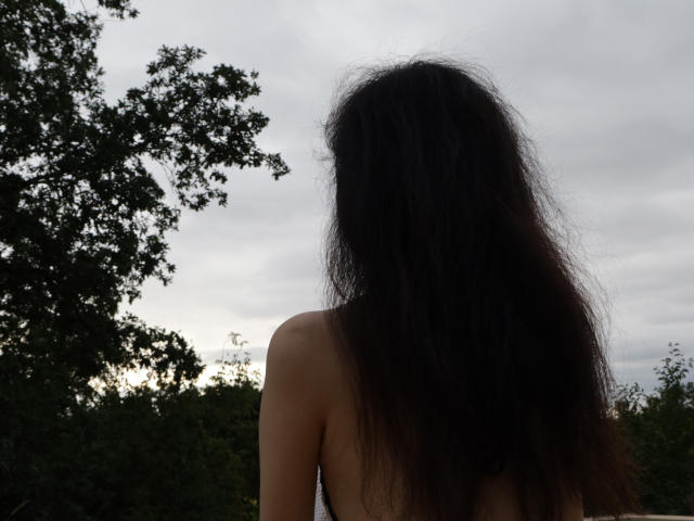 SheilaPass met lang, rommelig zwart haar, met de rug naar de camera, zonder top, waarbij haar blote schouders en bovenrug zichtbaar zijn. Bewolkte lucht, groene boom aan de linkerkant..