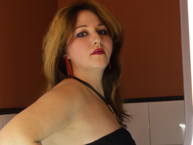 MirandaBrooks has a medium build with fair skin and shoulder-length brown hair. She wears a black halter top and red triangular earrings. Her makeup includes purple eyeliner and red lipstick. She looks slightly to the left with a relaxed expression. The lighting is warm, highlighting her facial features and upper body. The background is a brown and white wall. Her right arm is bent at the elbow.