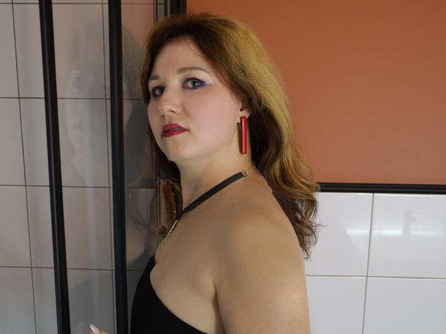 Miranda Brooks, met rode oorbellen, een zwarte haltertop en paars oogschaduw, kijkt opzij, met rode lippenstift en bruin golvend haar, in een kamer met tegels. Borsten zichtbaar..