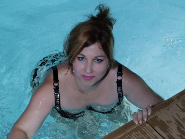 A photograph of MirandaBrooks in a swimming pool. She has fair skin, brown hair in a messy bun, and is wearing a black, strapless swimsuit with "FLY" in white letters. She's looking up at the camera with a slight smile, her arms resting on a wooden ladder. The water is clear, with light blue hues. Her breasts are visible, and she has a pink lip color. Her right hand has a silver ring. The texture of the water is slightly rippled.