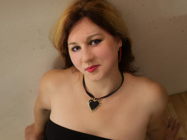 A close-up of MirandaBrooks with light skin and shoulder-length brown hair with blonde highlights. She wears a black strapless top, a black choker with a heart-shaped gold pendant, and red earrings. MirandaBrooks has dark eyeliner, pink lipstick, and a slight smile. Her right arm is visible, resting on a wooden surface. The image has a casual, slightly warm tone. The composition is centered, focusing on her upper body and face.