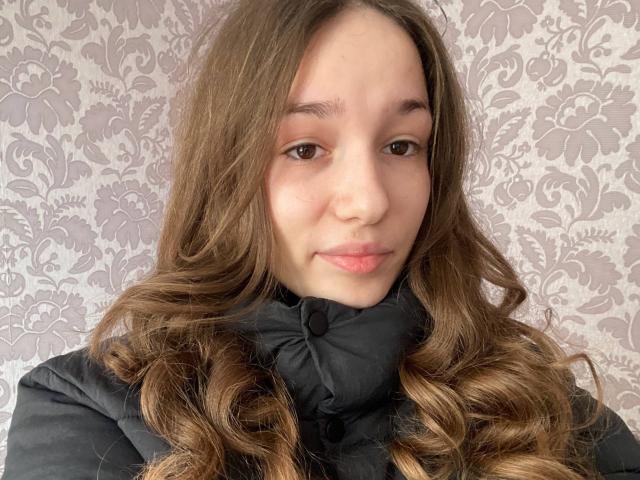 KaiaLein with wavy brown hair, light skin, and pink lips, wearing a black puffy jacket, has a subtle smile and brown eyes, looking directly at the camera.