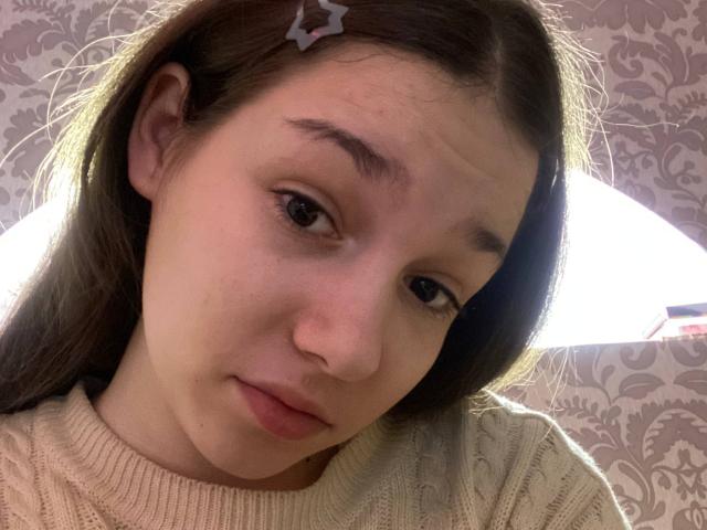 KaiaLein, a young woman with light brown skin and long brown hair, wears a beige, knitted sweater. She has a small star hairpin on the right side of her head. Her brown eyes are slightly downcast, and she has a subtle, relaxed expression. The image is close-up, focusing on her face and upper chest. Her eyebrows are thin, and her lips are closed. The lighting is soft, highlighting her facial features.