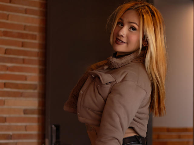 AzhunaStar with blonde hair, smiling, wearing a beige jacket, and a fur-lined scarf, looking over her shoulder, showing her tits slightly, in a dimly lit room.