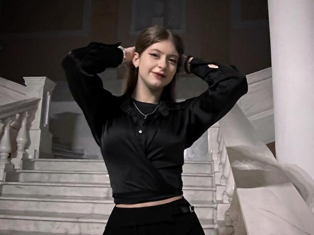 Palerianna with brown hair, wearing a black shiny long-sleeve top and black skirt, stands with hands behind head, smiling, showing small tits and a cute face.