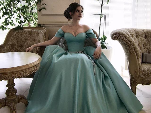Palerianna sitzt in einem prunkvollen Raum und trägt ein türkisfarbenes Ballkleid mit einem Schulterfrei-Schnitt, das ihre Brüste betont und ihre Taille formt. Ihr Gesichtsausdruck ist nachdenklich..