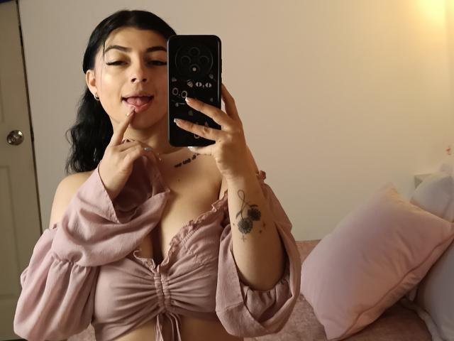 Anntonella Watson met zwart haar, tong uit, vinger op lip, roze top met één schouder, die de decolleté laat zien, tatoeages op de linkerarm, vasthoudend een telefoon, roze kussens op de achtergrond, kijkend in de spiegel..