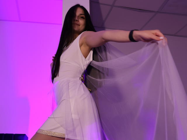 BabeRia with long brown hair, wearing a white dress, holding up a sheer fabric, purple and white light in the background, black wristband on her right arm.