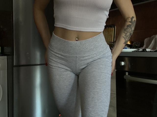 La camiseta blanca corta de Olieri, los pantalones grises muy ajustados, el piercing en el ombligo y el brazo tatuado, todo en una cocina con poca luz. Manos dentro de los pantalones. Esmalte de uñas rojo..