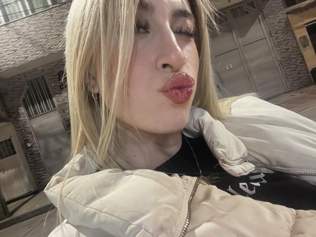 SammyViolet met blond haar, volle, roze lippen en gesloten ogen, gekleed in een beige gewatteerde jas, een zwart shirt en oorbellen, op een straat tijdens de nacht..