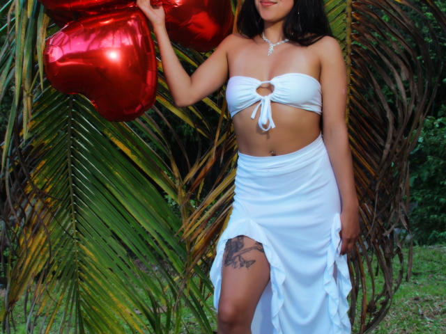 Lolisnoa with medium tits, white strapless top with keyhole, white skirt with thigh slit, holding red heart balloons, black tattoo on left thigh, standing in front of palm leaves.