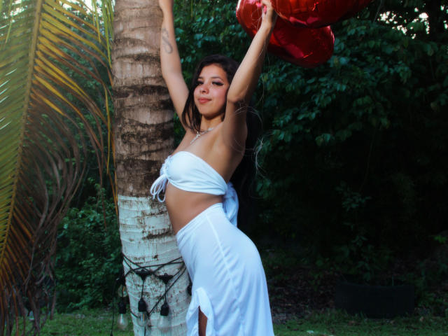 Lolisnoa with arms raised, wearing a white wrap-around dress, standing against a palm tree, holding a red shiny object, smiling, showing her tits and ass, in a green garden.