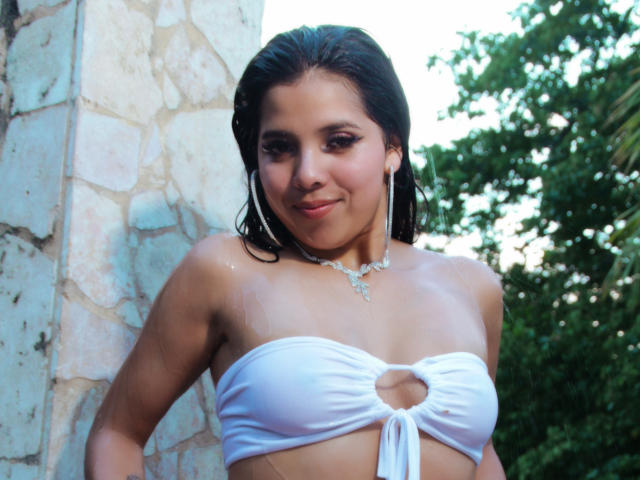 Lolisnoa with wet skin, wearing a white strapless top with a cutout, has dark hair, hoop earrings, and a necklace, smiling at the camera. Boobs are visible.