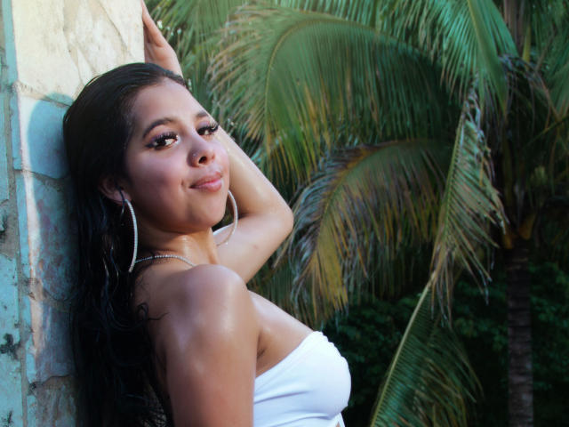Lolisnoa with dark hair, wearing a white strapless top, large hoop earrings, leaning against a wall, smiling, showing her ass, and palm trees in the background.