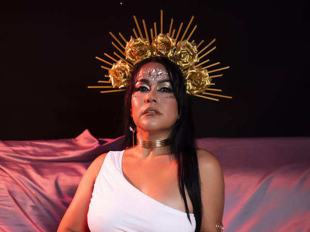 TamaraFoxy with dark hair, white one-shoulder top, gold rose crown, radiant face paint, and gold choker, sitting against purple fabric, looking seductive and confident.